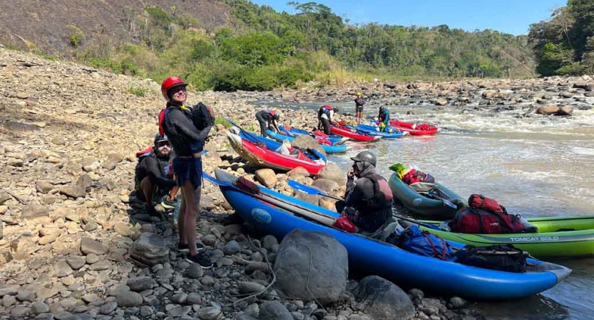 a group of watercraft rest on shore while a person in the foreground carries gear